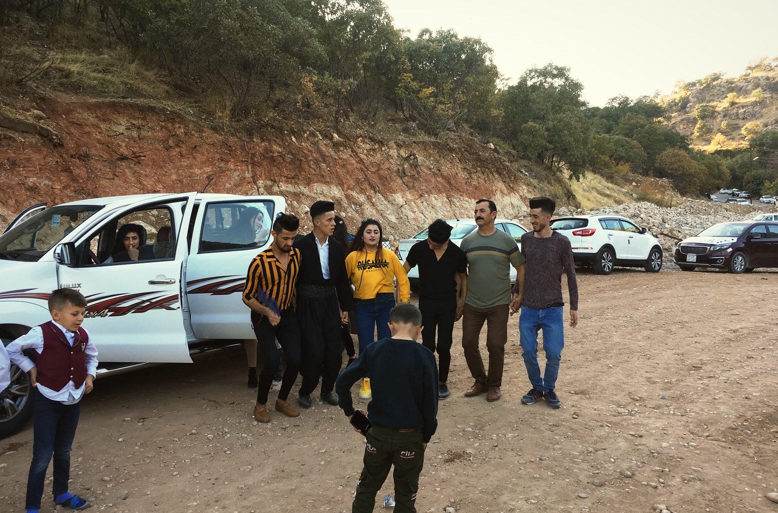 A line of young Yazidis dancing hand in hand beside parked cars on the track outside Lalish