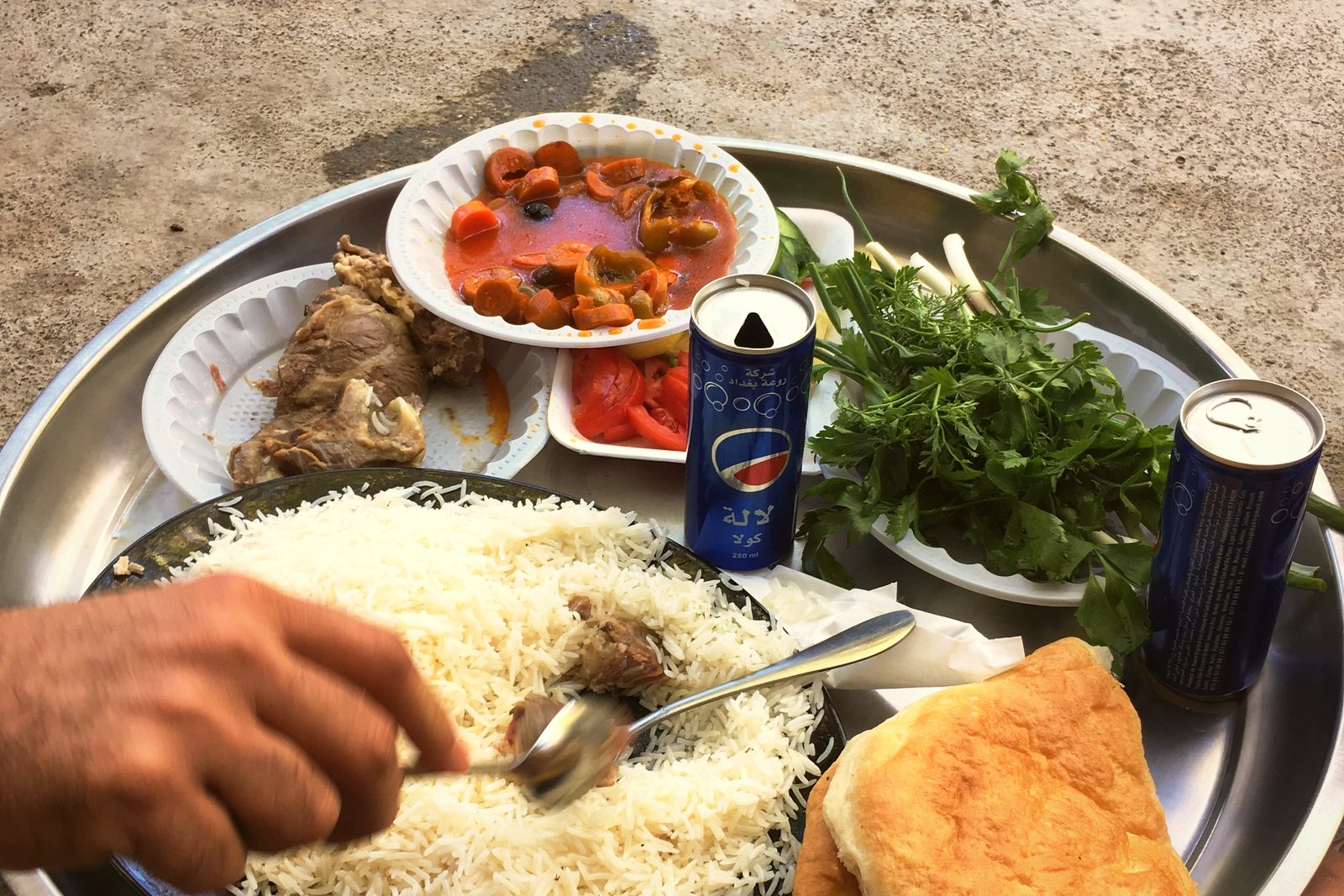 A large metal tray of rice, stew, salad, herbs and flatbread, with a hand reaching in with a spoon