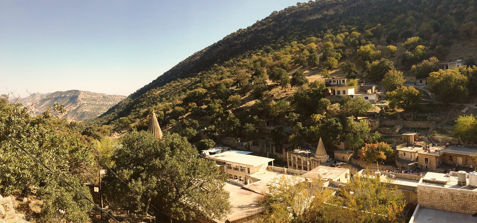The valley of Lalish seen from above — conical spires among trees, flat-roofed buildings and wooded mountains