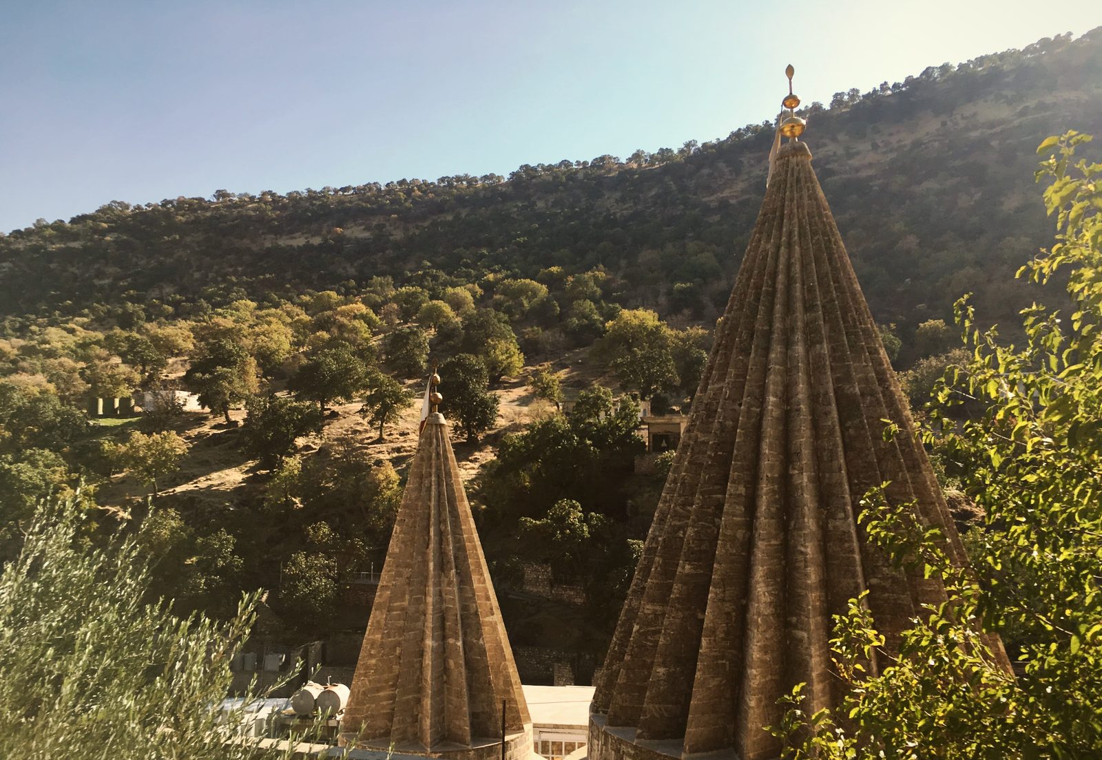 Two fluted conical spires of Lalish, the taller crowned with a gilded finial, against a wooded hillside
